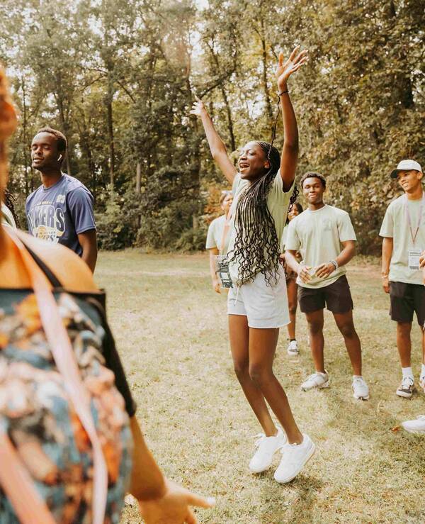 A young woman with long, braided hair jumps with arms raised in the air, surrounded by other students on a grassy area, with trees in the background. She wears a light green t-shirt, white shorts, and white sneakers. Other students wear light green or dark blue shirts.