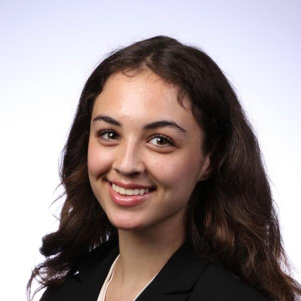 A young woman with dark wavy hair and a friendly smile wears a black blazer over a light shirt.