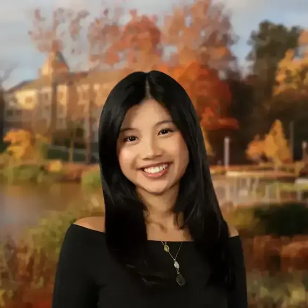 Smiling woman in black top and necklace, with Sorin Hall, colorful autumn trees, and a lake behind her.