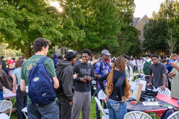 Diverse students smile and chat at an outdoor event, sunlight beaming through trees on Notre Dame campus.