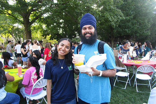 A smiling young woman and a man wearing a blue turban hold drinks and food outdoors.