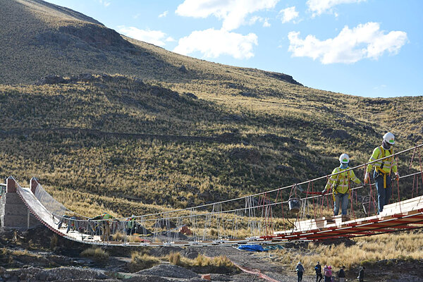 Two workers in yellow vests walk across a suspension bridge in a mountainous, grassy landscape.