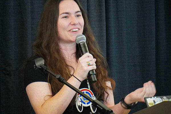 Woman speaks into a microphone wearing a black shirt with a beaded pendant and a tan skirt with black bear designs.
